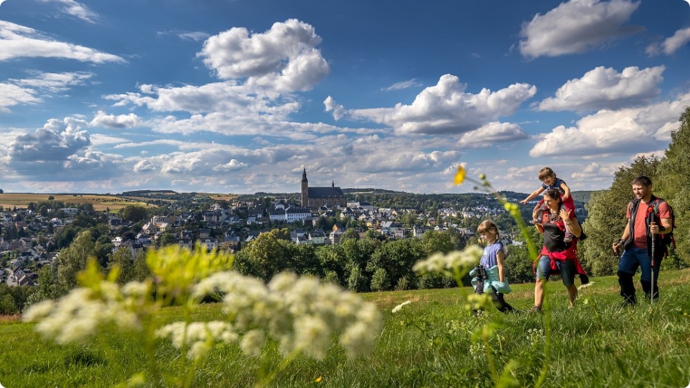 Eine Familie wandert in der Natur über eine Wiese, im Hintergrund eine Stadt und blauer Himmel mit Wolken.
