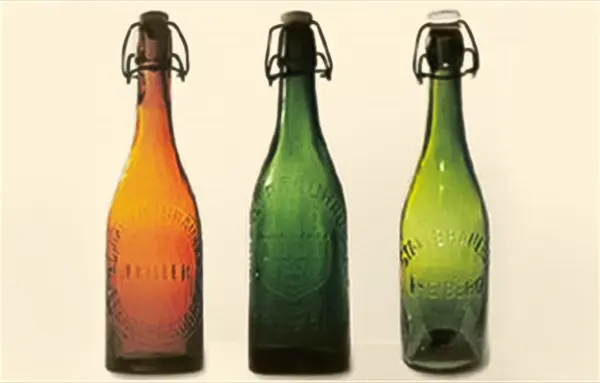 Drei farbige Glasflaschen in Orange, Grün und Gelb stehen nebeneinander. Jede Flasche hat einen Bügelverschluss.