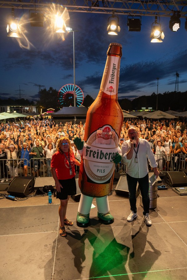2 Personen stehen mit einem übergroßen Flaschenmaskottchen auf einer Bühne, dahinter jubelndes Publikum.