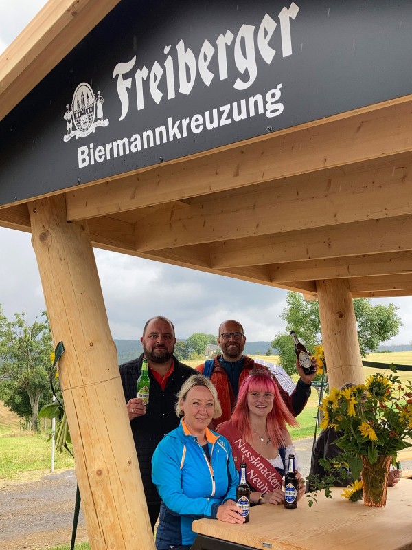 Eine Gruppe von Menschen mit vier Flaschen Freiberger Bier rasten in einer neu aufgebauten Rasthütte mit der Aufschrift Freiberger Biermannkreuzung.
