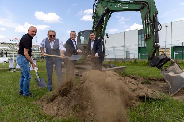 Vier Personen graben auf einer Wiese mit vier Spaten, dahinter steht ein Bagger und ein Freiberger Schrirm.