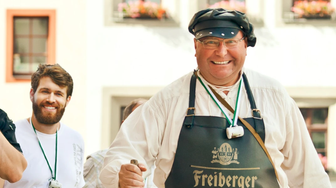 Ein Mann in mittleren Jahren lächelt, trägt eine Schürze und eine traditionelle Kopfbedeckung.