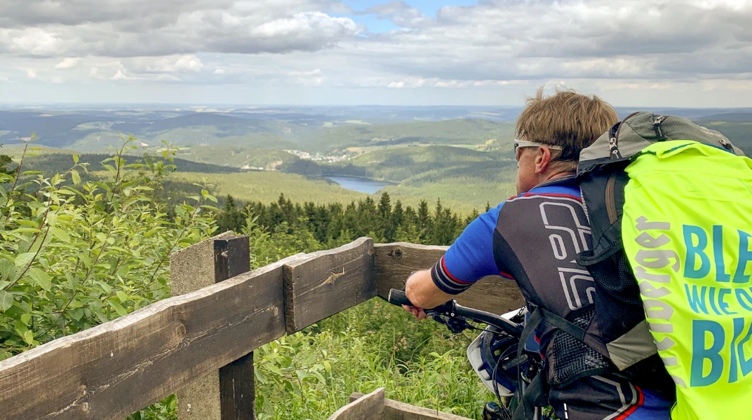 Ein Radfahrer betrachtet von einem Holzzaun aus eine hügelige Waldlandschaft mit Seen.