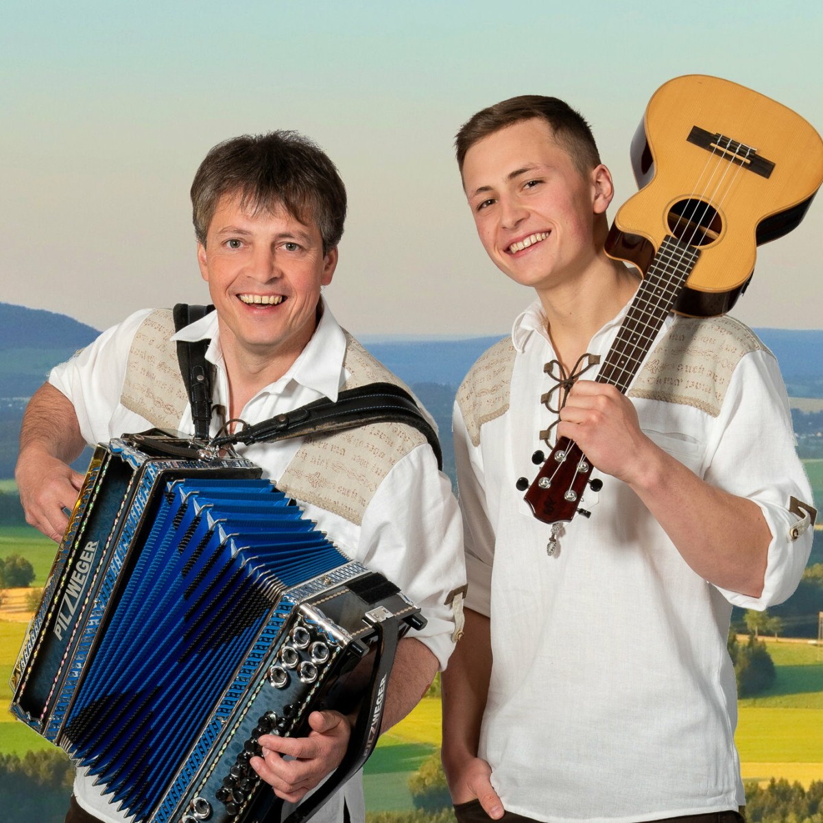 Zwei Musiker im traditionellen Outfit mit Akkordeon und Gitarre vor ländlicher Kulisse.