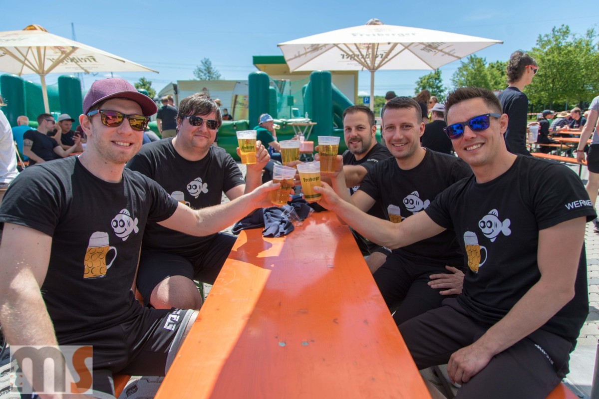 Fünf Männer sitzen an einem orangefarbenen Tisch und stoßen mit Biergläsern an.