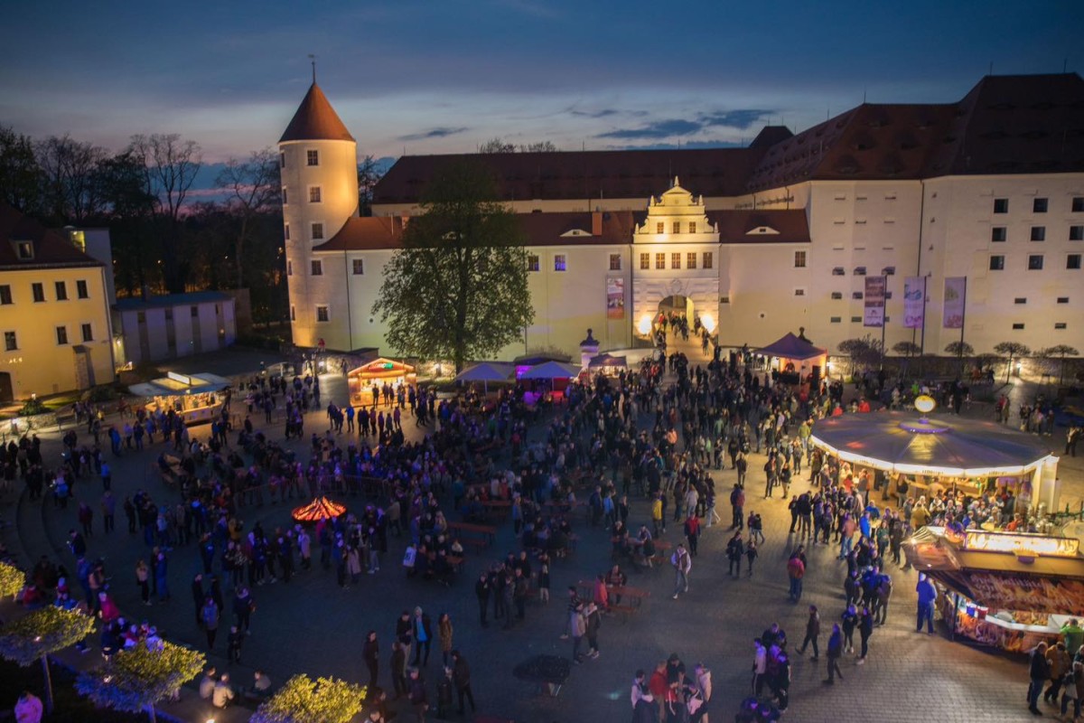 Blick von oben auf ein Fest, Personen sind umringt von Essensbuden und einem Karusell, in der Mitte ist ein Lagerfeuer. Im Hintergrund ist ein Schloss zu sehen.