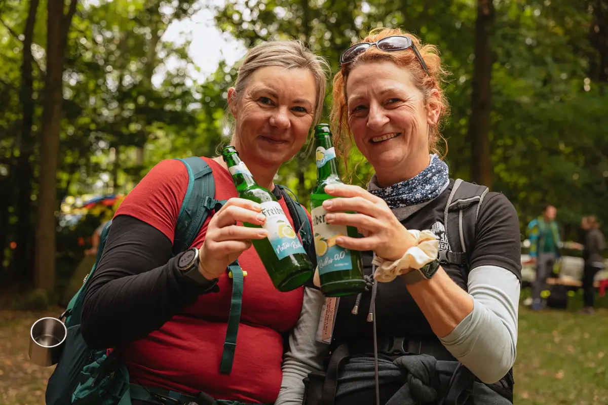 Zwei Frauen stoßen mit Freiberger Bierflaschen an und tragen Wanderrucksäcke. Im Hintergrund Bäume und Personen im Wald.
