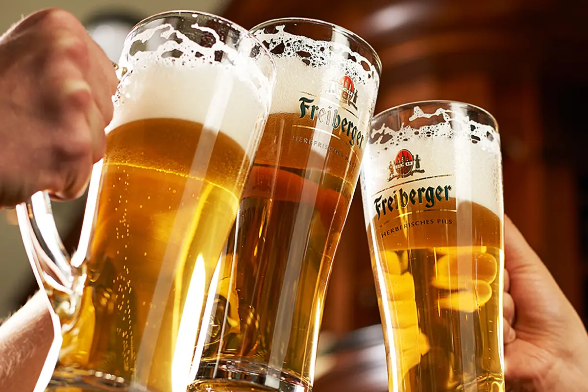 Drei Bierglaeser werden von Händen zum Anstoßen zusammengefuehrt. Holzbar im unscharfen Hintergrund.