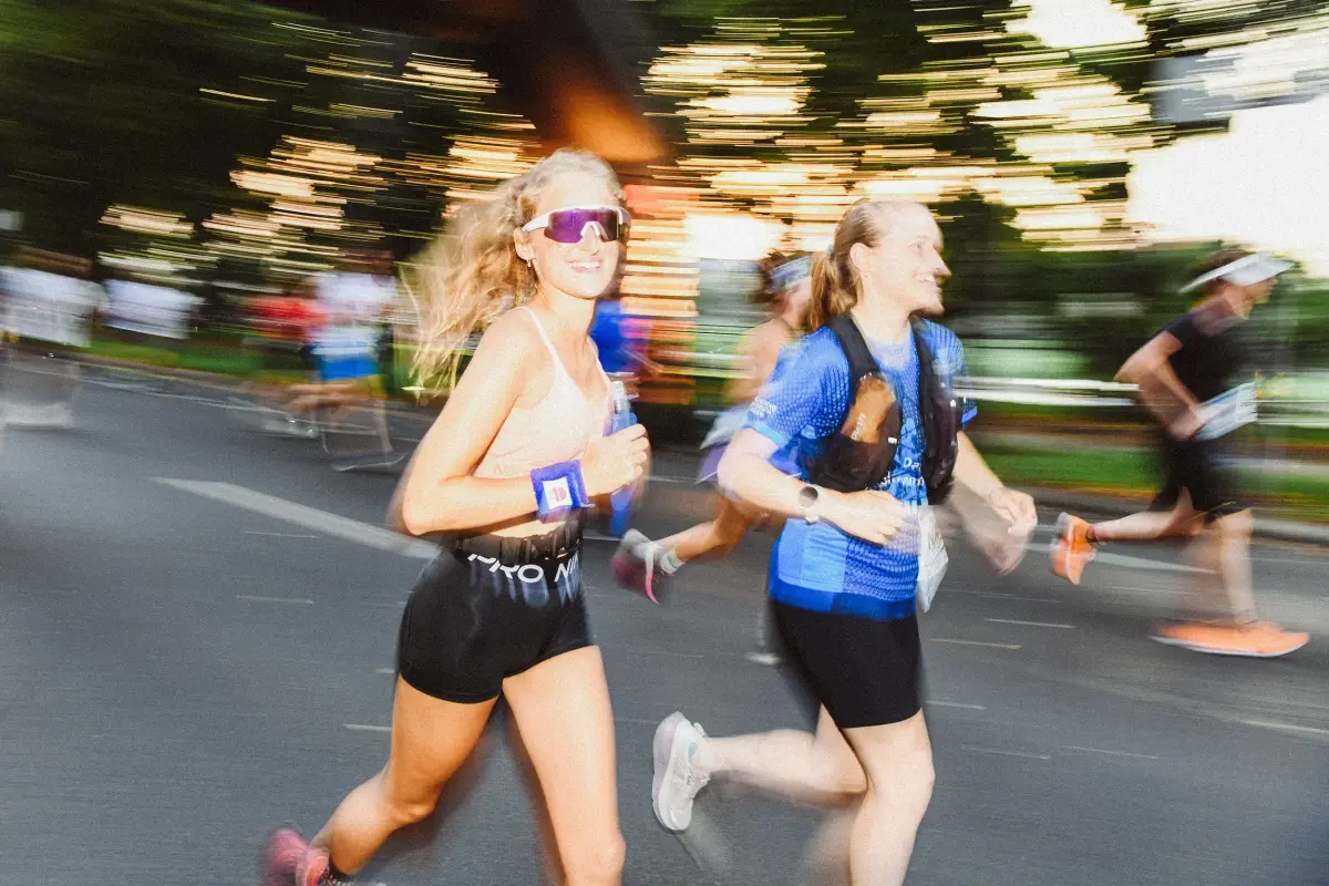 Zwei Frauen laufen nebeneinander bei einem Straßenlauf. Im Hintergrund weitere Läufer auf Straße und verschwommene Bäume.