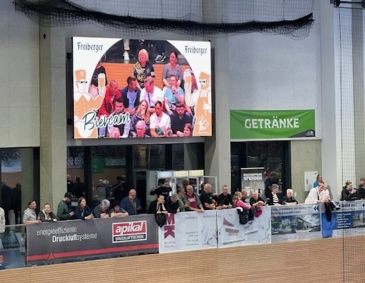 Mehrere Personen stehen am Spielfeldrand in einer Sporthalle, darüber ein großes Display mit Freiberger Werbung.