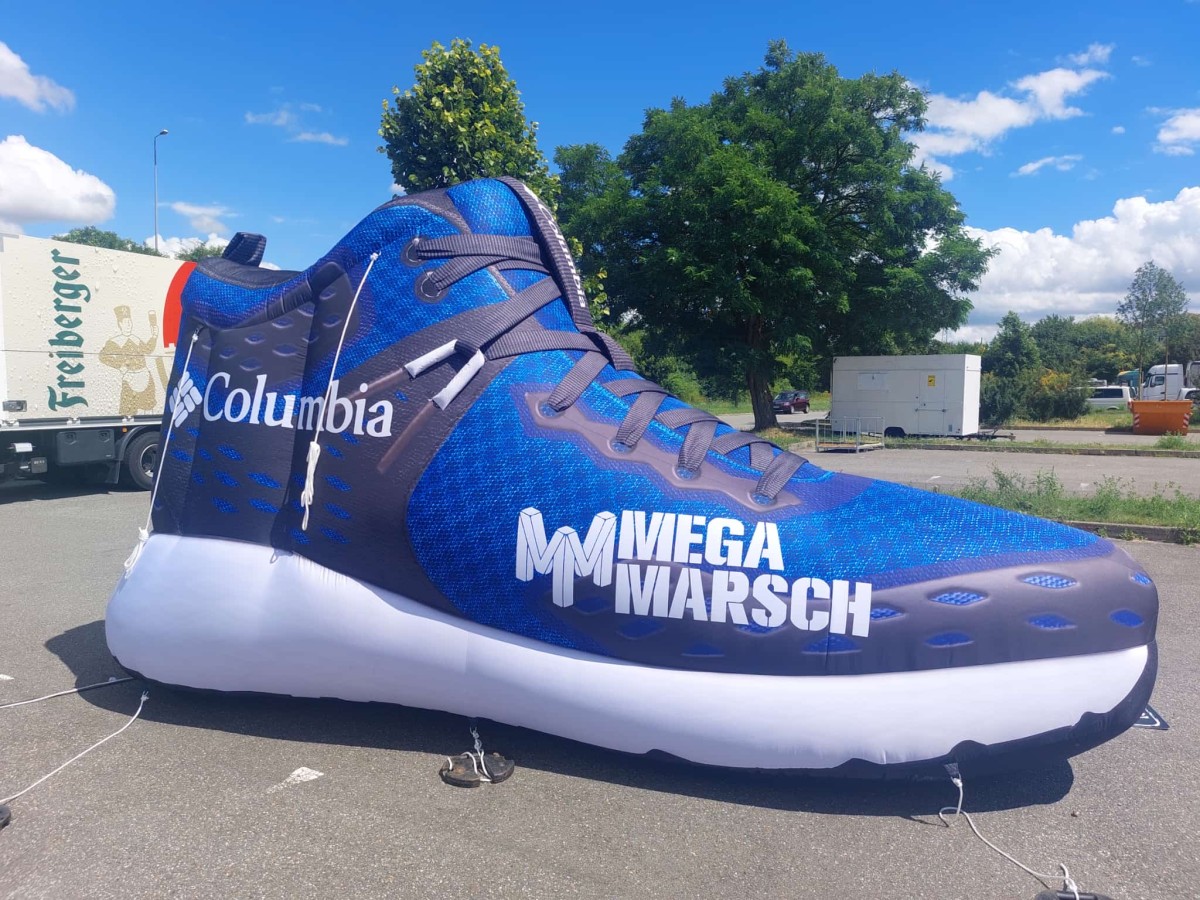 Ein rieseger aufgeblasener blauer Turnschuh mit der Aufschrift Mega Marsch steht auf einer Asphaltfläche, im Hintergrund ein Freiberger LKW.