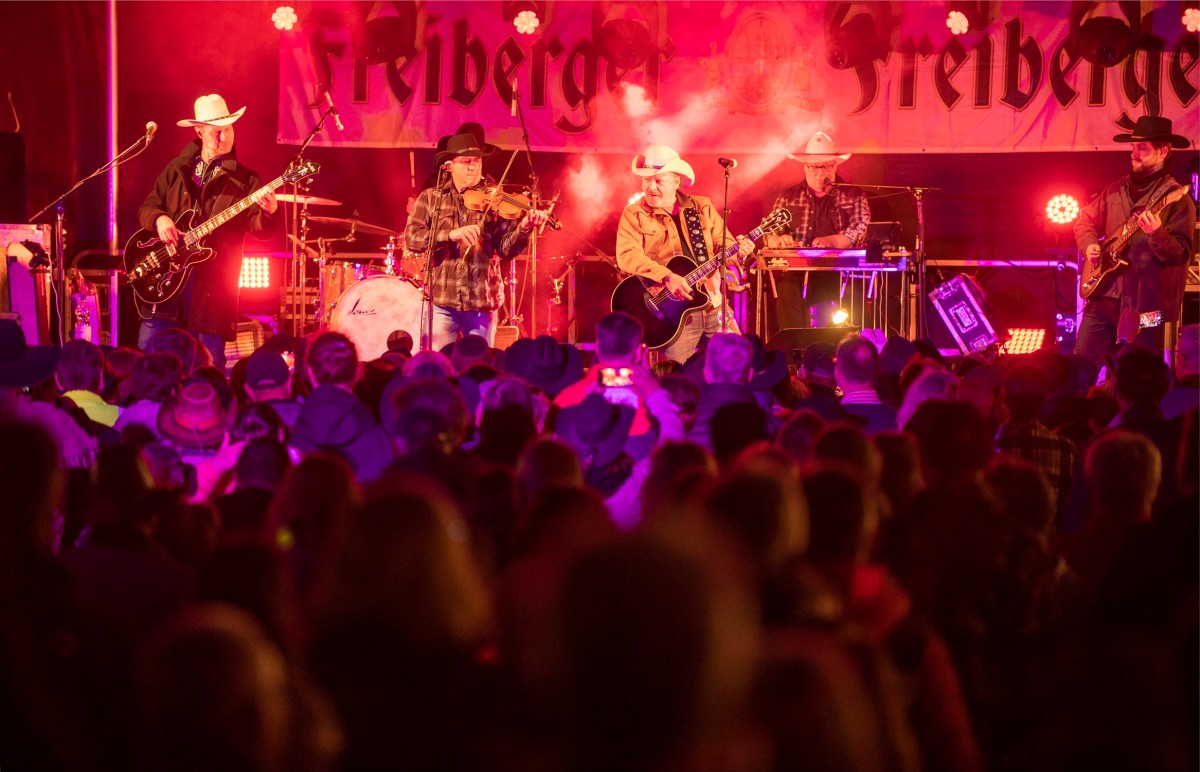 Eine Band spielt auf einer Bühne Musik. Vor der Bühne steht ein Publikum.