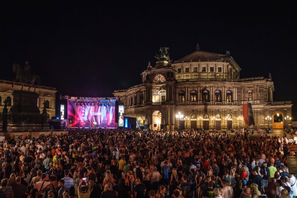 Vor einem großen, alten Gebäude steht eine Bühne. Vor ihr sind zahlreiche Menschen versammelt.