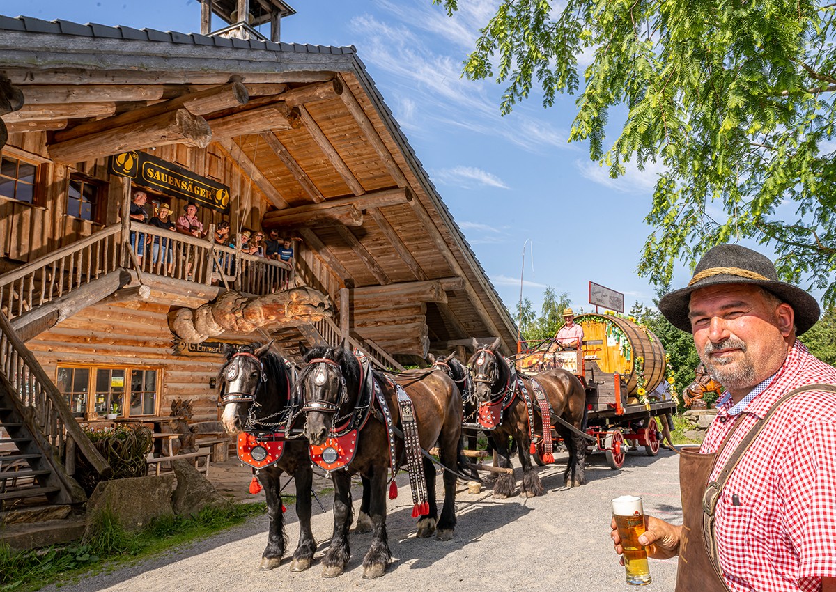 Ein Mann mit einem Glas Freiberger Bier steht vor einer Almhütte neben einer Bierkutsche mit 4 Pferden.