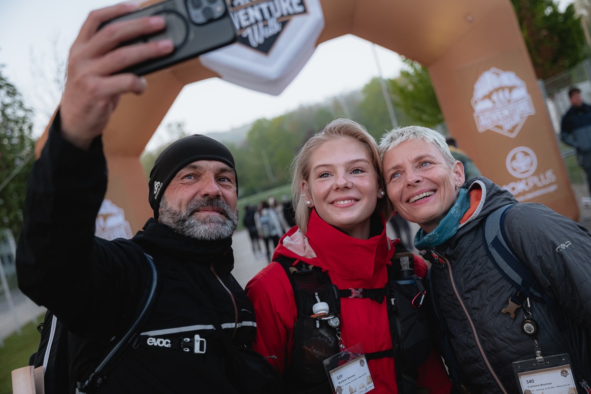 Drei Menschen mit Outdoorkleidung stehen vor dem Adventure Walk Zieleinlauf und machen ein Selfie.