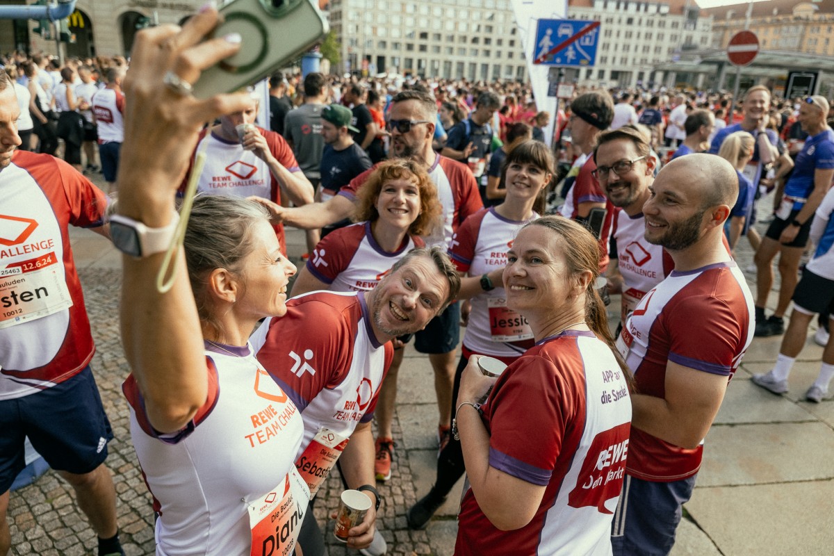 Ein Team aus zehn Menschen in Sportkleidung steht in einer Menge von anderen Sportlern auf einem großen Platz, sie machen ein Selfie.