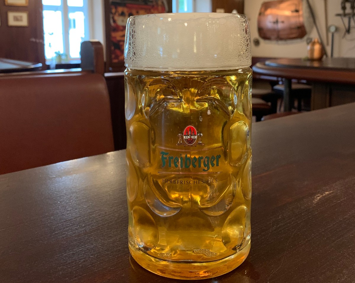 Ein Freiberger Pils Maßkrug steht auf einem braunen Tresen im Hintergrund Tische und Fenster.