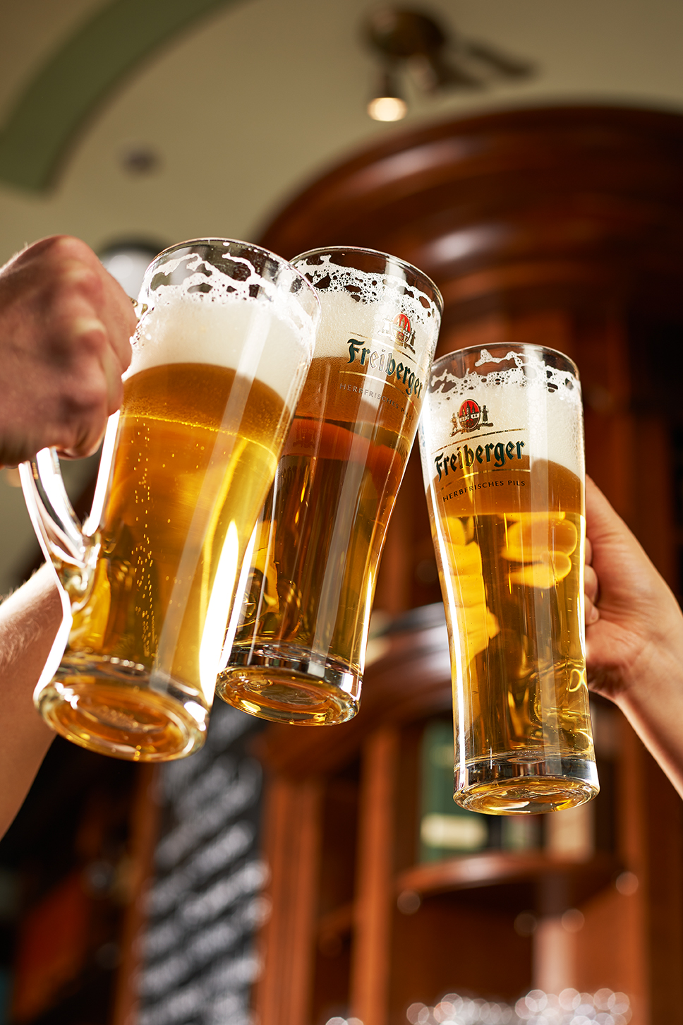Drei Hände stoßen mit frisch gezapftem Bier an, zwei davon mit Freiberger Pils im Markenglas.