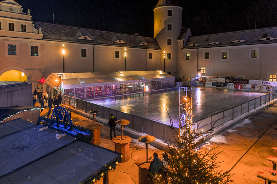 Schloss Freudenstein bei Nacht: beleuchtete Eisbahn im Innenhof mit Rundturm, Glaspavillon, Stehtischen und Weihnachtsbaum.