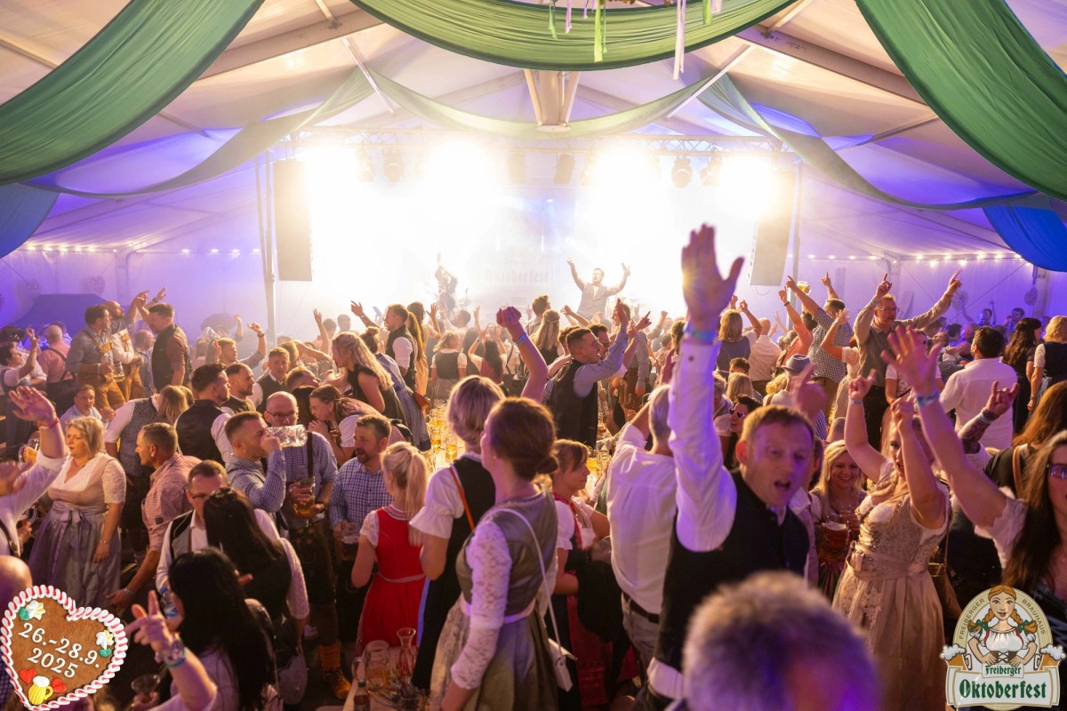 Menschen feiern auf dem Freiberger Oktoberfest in einem Festzelt, im HIntegrund spielt eine Band auf der Bühne.