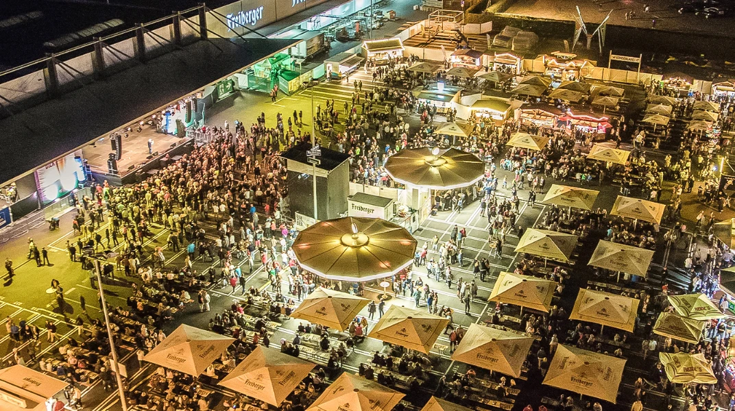 Eine nächtliche Ansicht zeigt eine belebte Außenveranstaltung mit vielen Menschen und Marktständen.