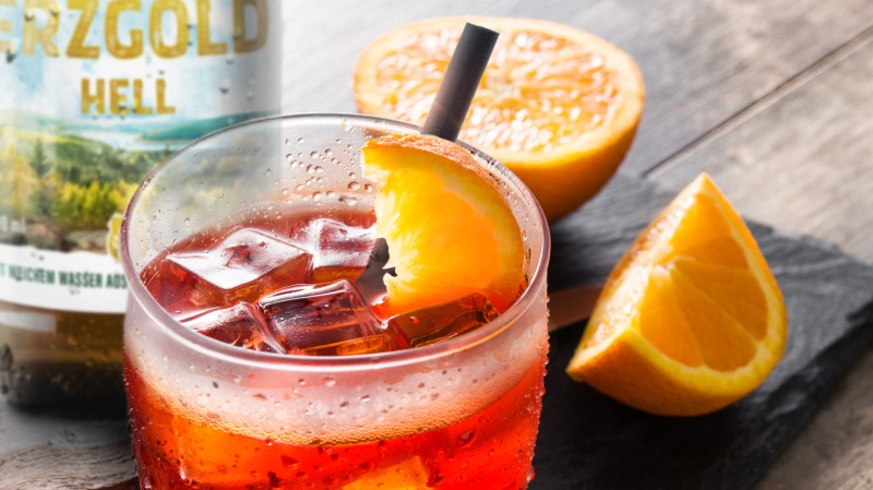 Ein Glas mit roter, kohlensäurehaltiger Flüssigkeit und Eiswürfeln ist abgebildet, garniert mit einer Orangenscheibe