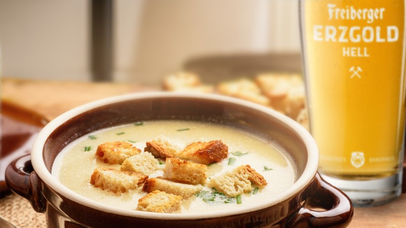 Ein Suppenteller gefüllt mit einer Bier-Käsesuppe, garniert mit Schnittlauch und Croûtons. Daneben ein Glas Freiberger Erzgold Hell.