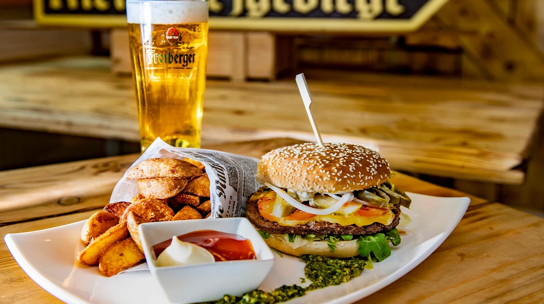 Ein Hamburger mit Beilagen und ein großes Bier stehen auf einem Holztisch.