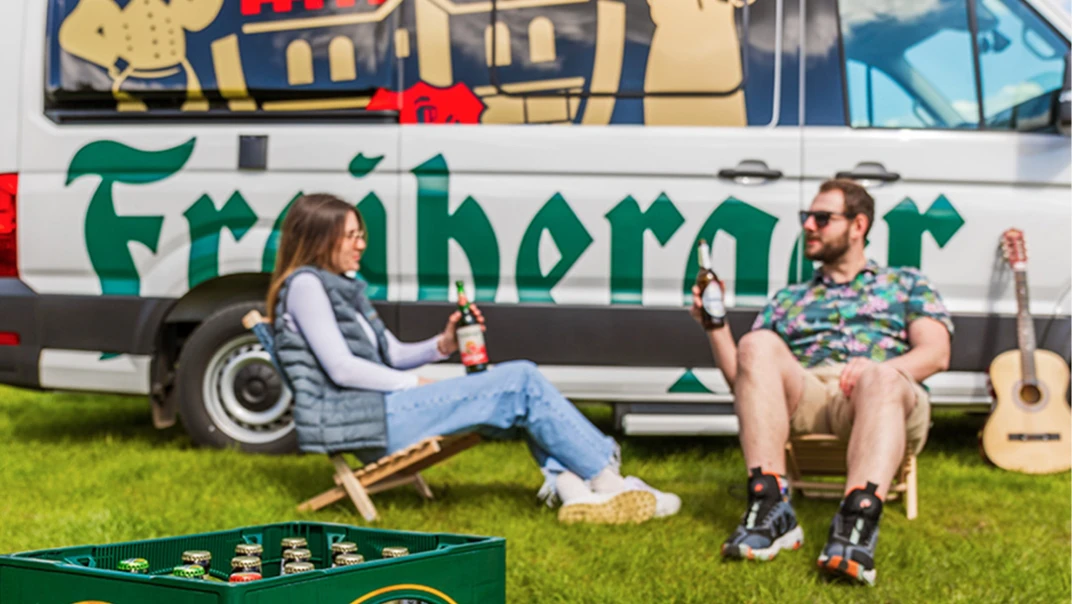 Zwei Personen sitzen entspannt auf Campingstühlen und halten Getränkeflaschen neben einem bunten Van.