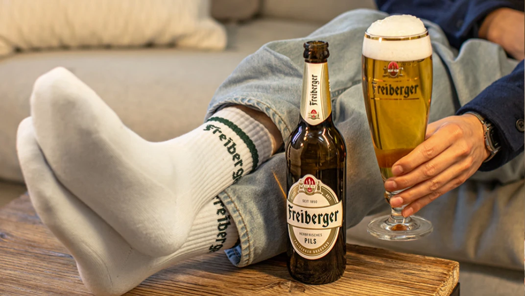 Ein Mann sitzt mit weißen Socken und hochgekrempelten Jeans entspannt auf einem Sofa. Neben ihm steht eine Flasche und ein Glas gefüllt mit goldener Flüssigkeit.