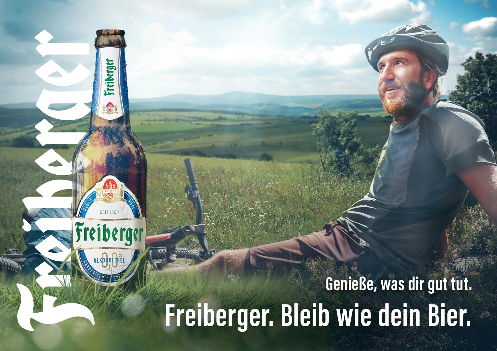 Ein Mann mit Fahrradhelm sitzt im Gras und schaut über eine hügelige Landschaft.