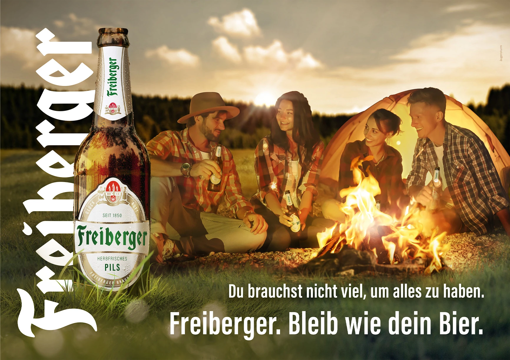 Vier Personen sitzen abends lächelnd an einem Lagerfeuer und halten Getränke.