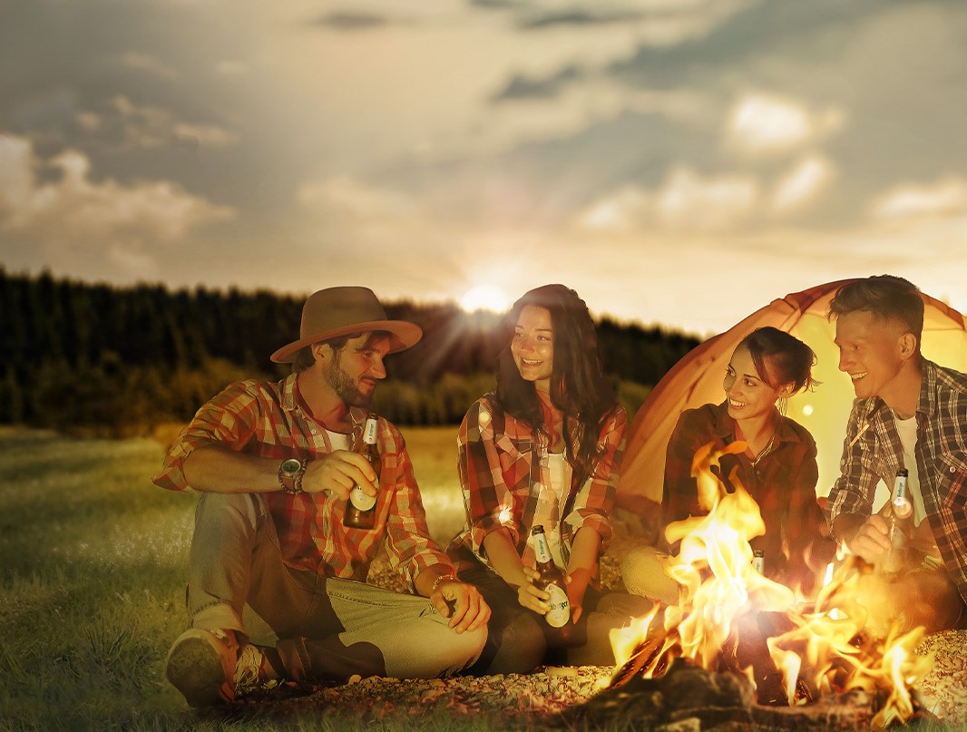 Vier Freunde sitzen lachend um ein Lagerfeuer in einer abendlichen Naturkulisse.